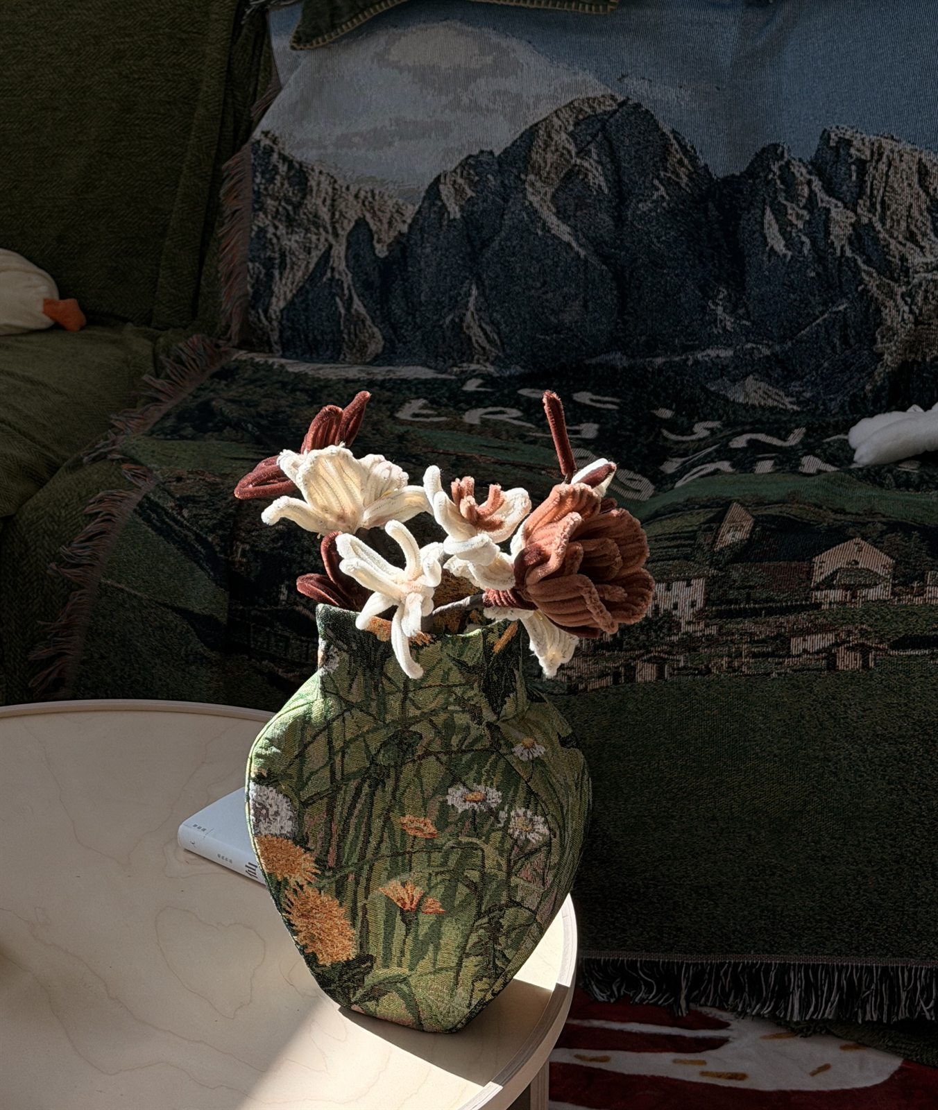 Ceramic floral vase on a table in front of a mountain tapestry draped over a green sofa